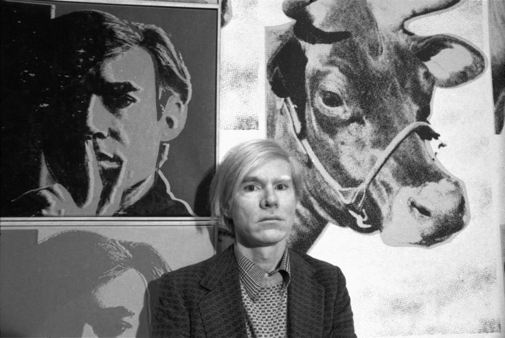 Andy Warhol sitting next to his Self Portrait artwork.