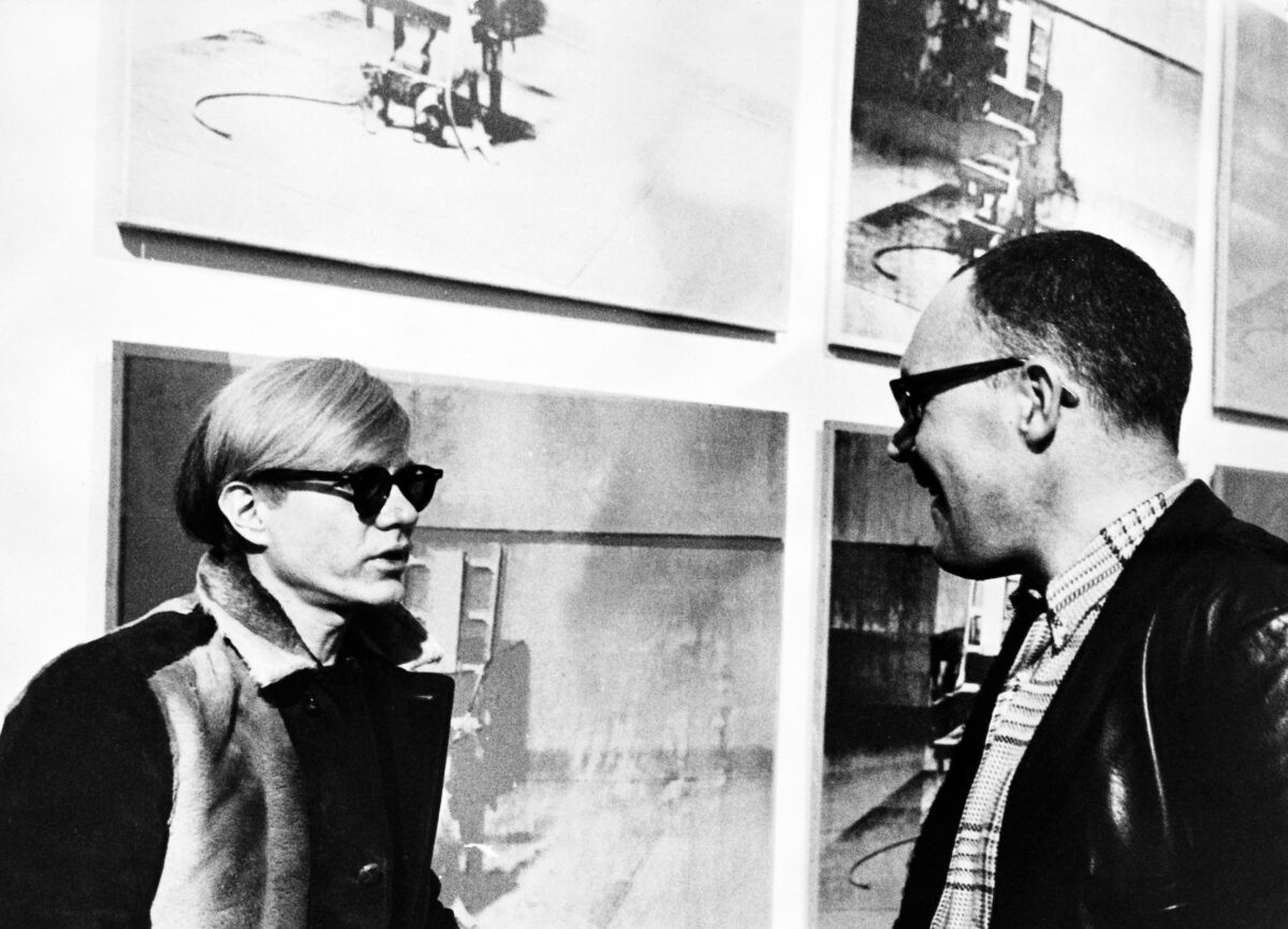 Andy Warhol standing in front of his Electric Chairs screen prints.