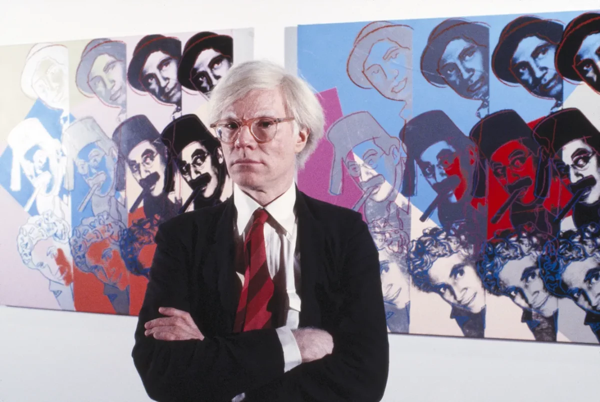 Andy Warhol standing in front of two of his Marx Brothers screenprints.