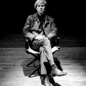 Black and white photo of Andy Warhol sitting in a director's chair with legs crossed, by Marshall Swerman (signed photograph).
