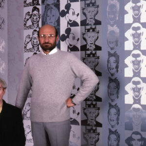 Ronald Feldman standing next to Andy Warhol, sitting down, in front of Warhol artworks.