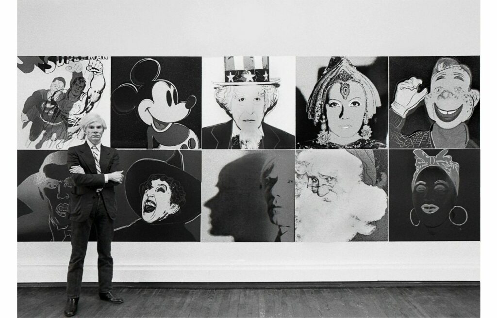 Andy Warhol stands in front of his screenprints from the Myths series, hanging on the wall behind him.