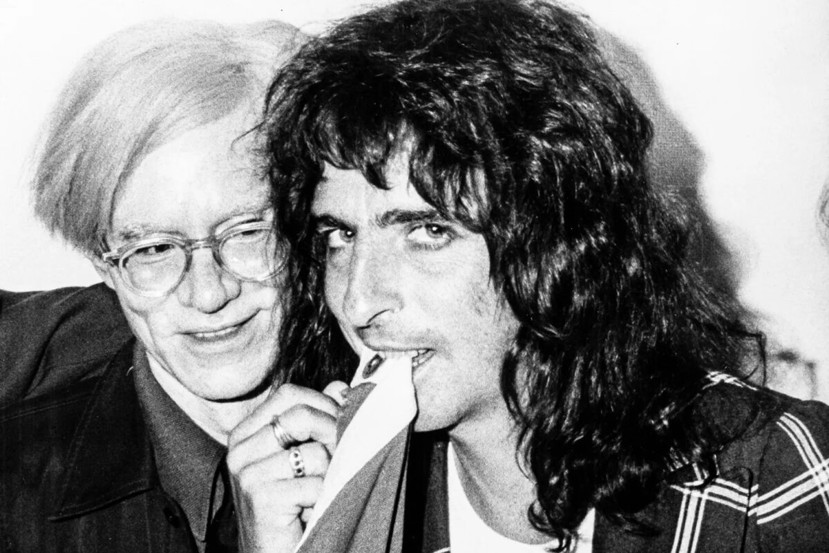 Alice Cooper looks at the camera and bites a piece of fabric while Warhol, behind him, smiles and looks at the fabric.