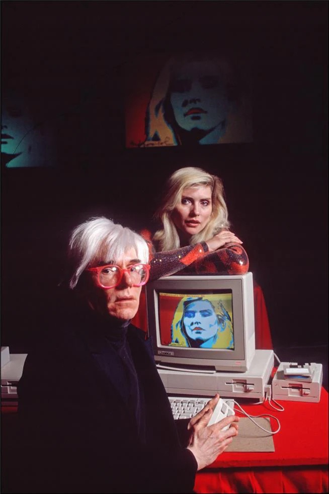 Andy Warhol and Debbie Harry sitting at a computer with a digital art piece of Debby Harry by Warhol on the screen.