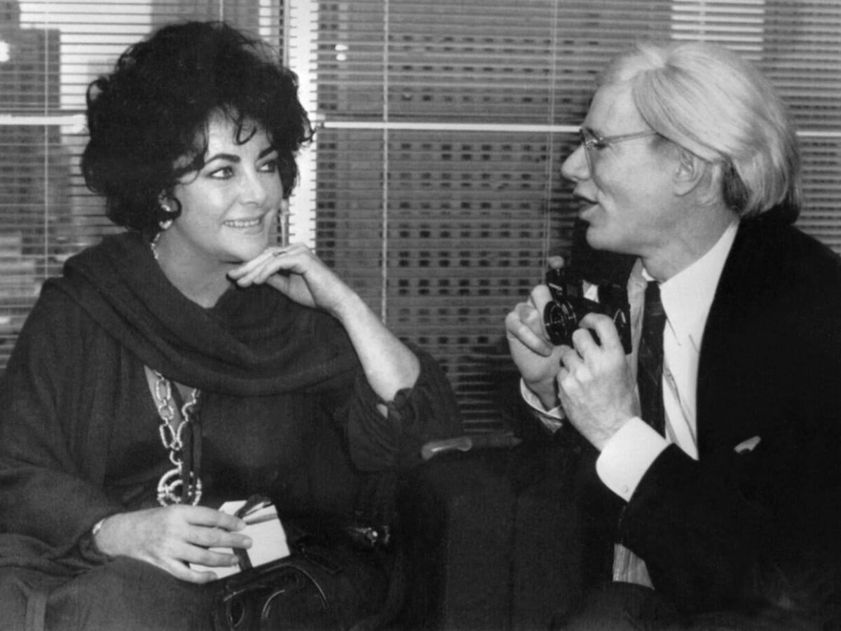 Elizabeth Taylor and Andy Warhol talking as Warhol holds a camera and Taylor poses for him.