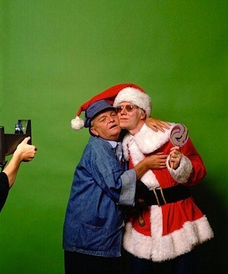 Truman Capote and Andy Warhol pose for a photo together in Christmas attire against a green backdrop.