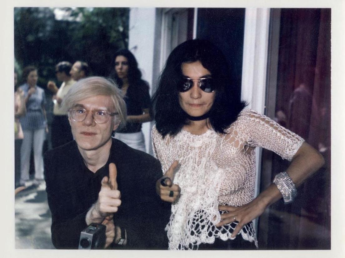 Yoko Ono and Andy Warhol pose while doing "finger guns" at the camera outside.